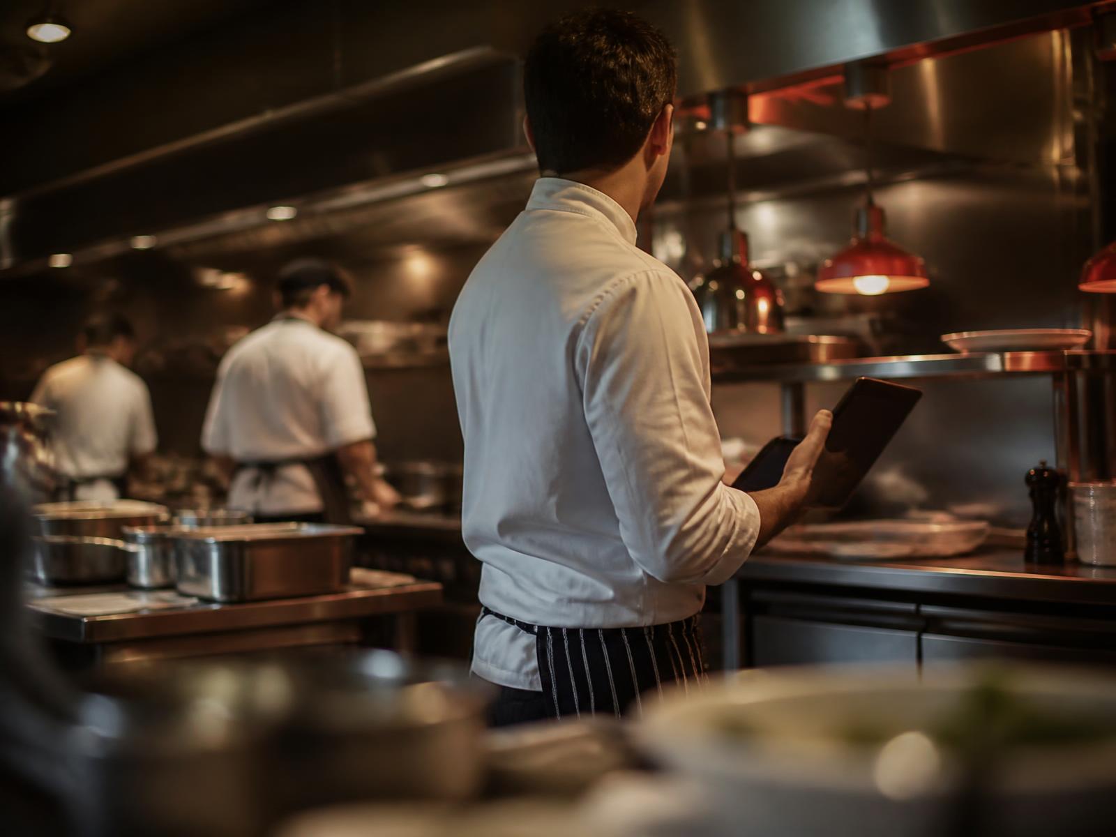 Chef reviewing digital food safety checks on a tablet in a busy commercial kitchen